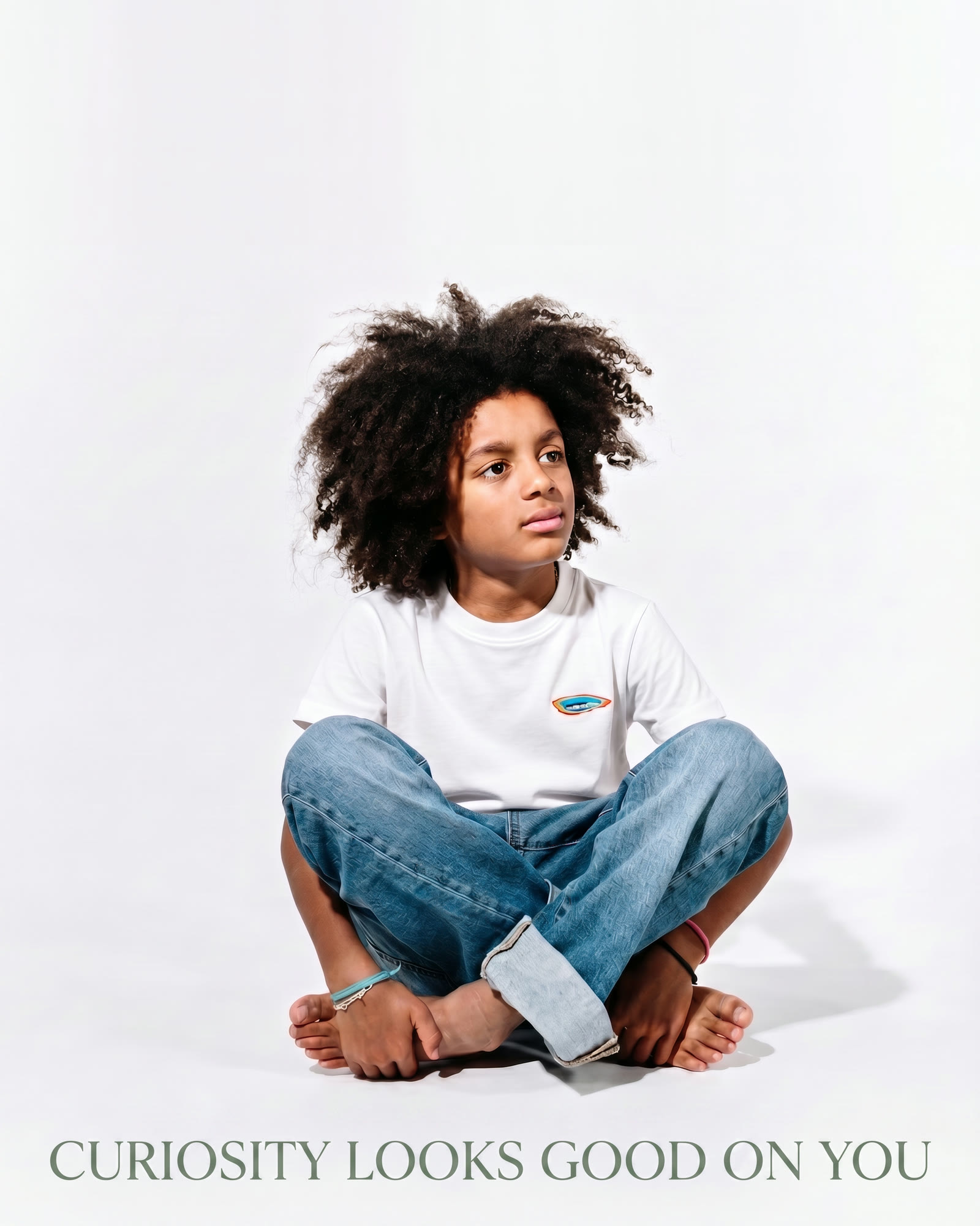 A boy sitting cross-legged in a white studio wearing the Enlightened Generation tee, above the line 'Curiosity looks good on you'